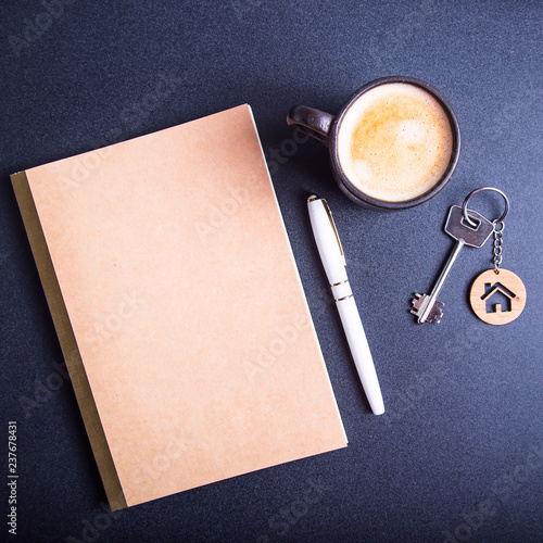 The keys to the new apartment with a wooden key chain house on a black table next to the crafting pad and a cup of coffee. Buying an apartment or house concept. Top view, flat lay