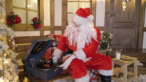 Santa Claus sits near the house between christmas trees, drinks milk, eats cookies, listens christmas songs on vinyl by gramophone, relaxes, enjoys, miss alone