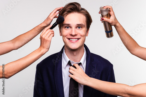 Are you ready for a date? Handsome young man surrounded by hands holding grooming tools looking at camera with smile while standing against white background
