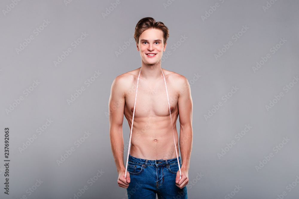 In perfect shape. Handsome shirtless young man in jeans holding measuring tape and looking at camera with smile while standing against grey background