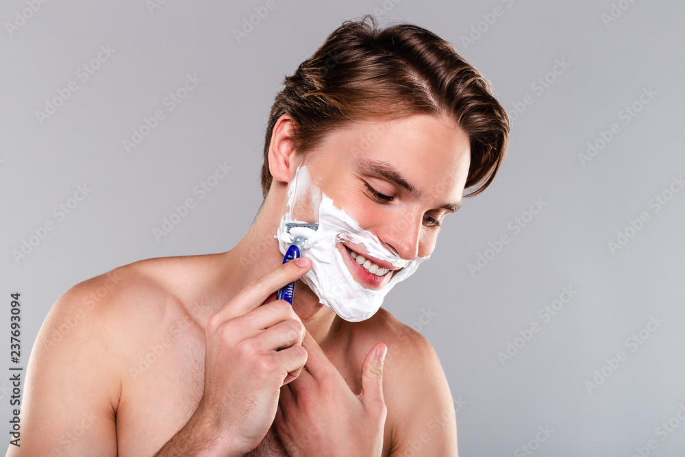 Shaving with no pain. Portrait of handsome shirtless young man shaving his beard with smile while standing against grey background