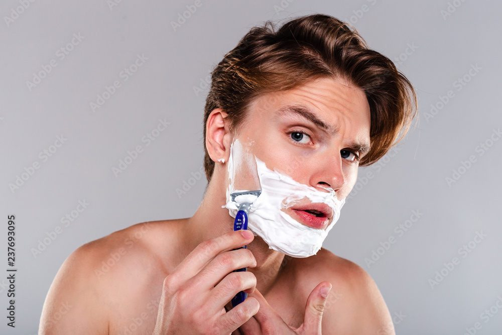 Clean and handsome. Portrait of handsome shirtless young man shaving his beard and looking at camera while standing against grey background