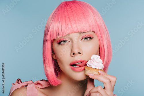 fashionable girl in pink wig licking buttercream from cupcake isolated on blue