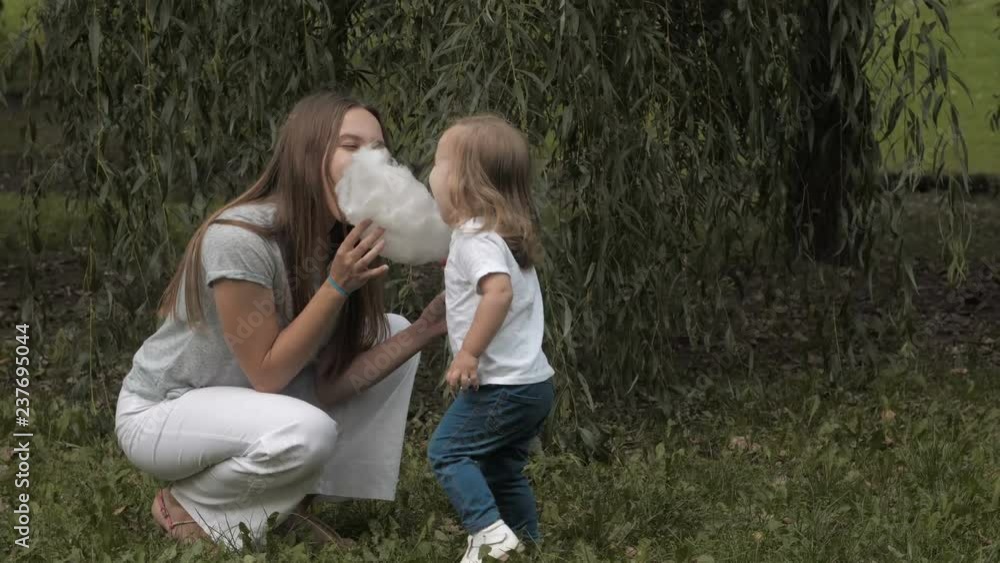 Doughter with mom in the park having fun and sharing cotton candy. Portrait of smiling baby eats cotton candy and looking at the camera. Doughter with mom in the park having fun and sharing cotton candy. Portrait of smiling baby eats cotton candy and looking at the camera.