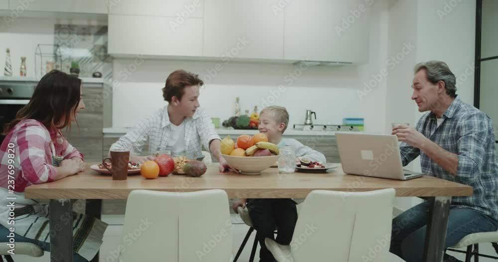 Charismatic mature family eating healthy food, they have a breakfast sitting at the dinner table happy spending a time together , in a modern kitchen , father working on the laptop while drinking his Charismatic mature family eating healthy food, they have a breakfast sitting at the dinner table happy spending a time together , in a modern kitchen , father working on the laptop while drinking his