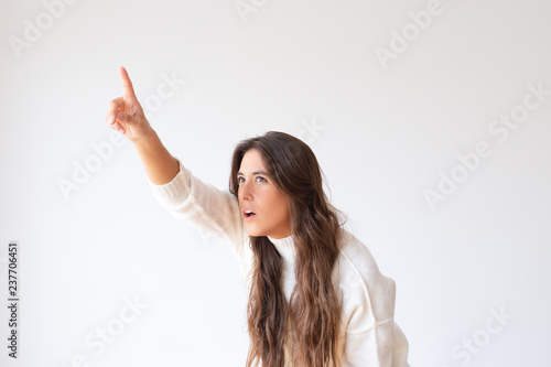 Amazed young woman pointing at something and looking up. Shocked girl in white sweater showing discount with finger. Isolated on white. Sale concept