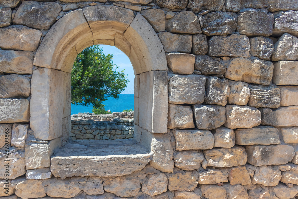 wall with a window in the ruins of the ancient city with a view of the sea, a tree and people, selective focus