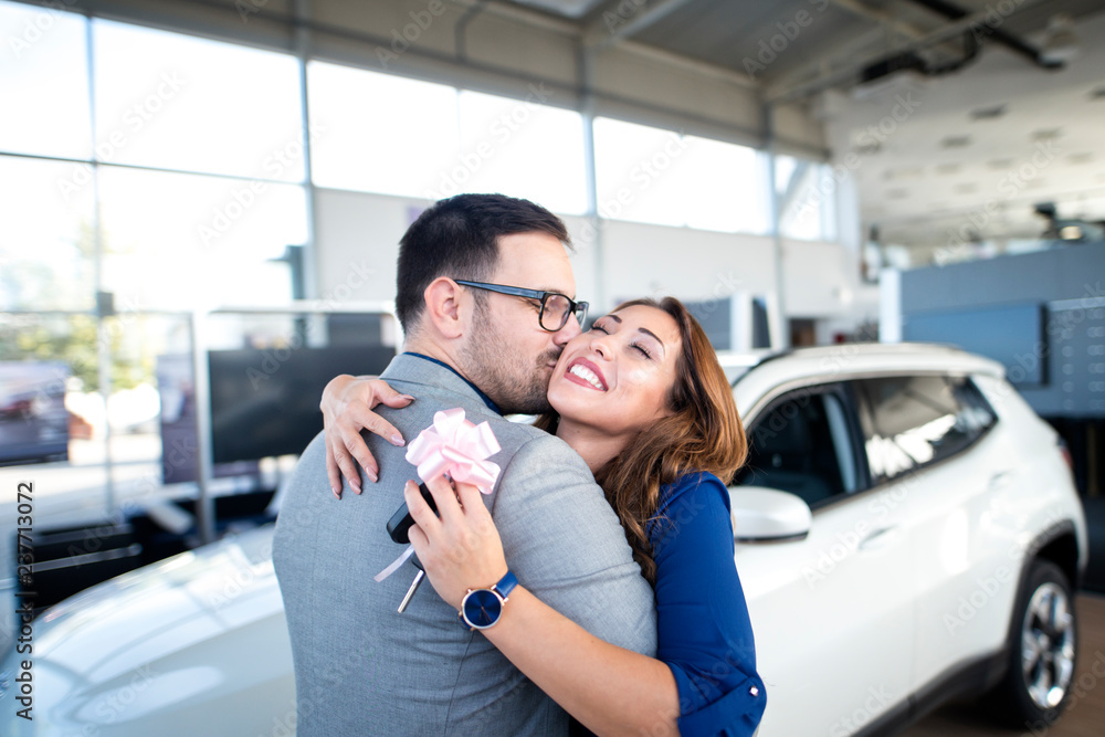 Young happy couple celebrating purchase of their new car at vehicle dealership. Woman holding car keys while man is kissing her. Young happy couple celebrating purchase of their new car at vehicle dealership. Woman holding car keys while man is kissing her.
