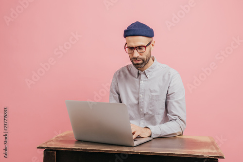 Concentrated hipster student reads information and checks data, focused in monitor of laptop computer, keyboards something, sits at desk, wears formal clothes, isolated over pink studio wall