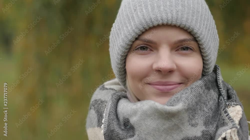 Portrait of young adorable woman in grey hat, scarf, blush jacket, standing on the wind, enjoying cold autumn day, smiling and looking into camera with fall colors background. Extreme close up shot. Portrait of young adorable woman in grey hat, scarf, blush jacket, standing on the wind, enjoying cold autumn day, smiling and looking into camera with fall colors background. Extreme close up shot.