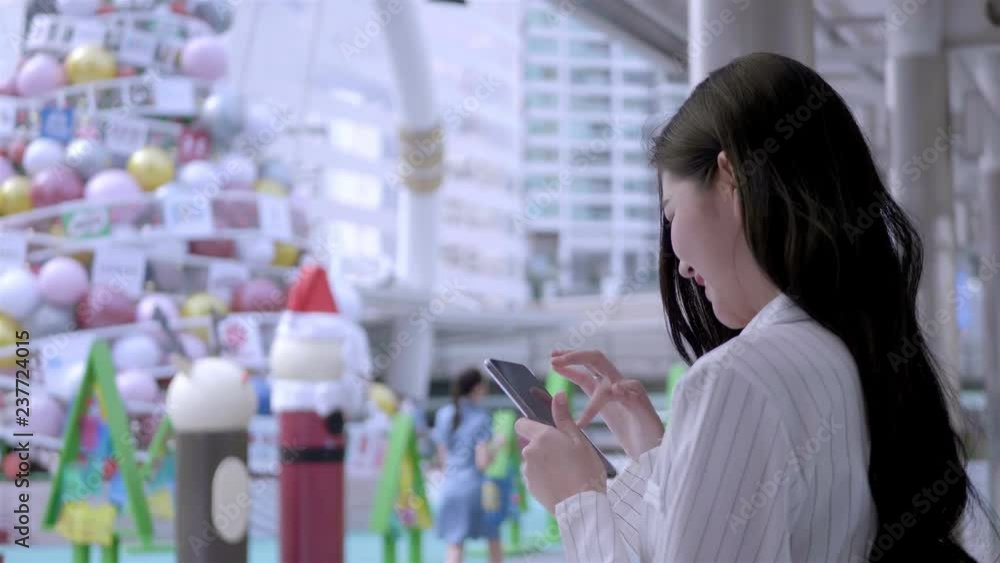 Young Asian businesswoman Using Smart Phone and smiling happily in city centre Thailand. communication, everyday life Technology Concept. SLOW MOTION Young Asian businesswoman Using Smart Phone and smiling happily in city centre Thailand. communication, everyday life Technology Concept. SLOW MOTION