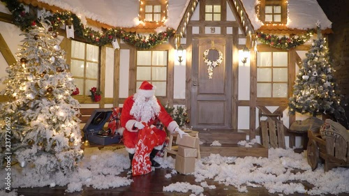 Santa Claus is packing gifts or presents in bag, sits outside the house between christmas trees, drinks milk, listens christmas songs on vinyl by gramophone, preparing for Christmas