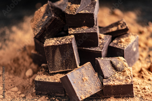 Pieces of dark bitter chocolate with cocoa powder on dark wooden background. Concept of confectionery ingredients