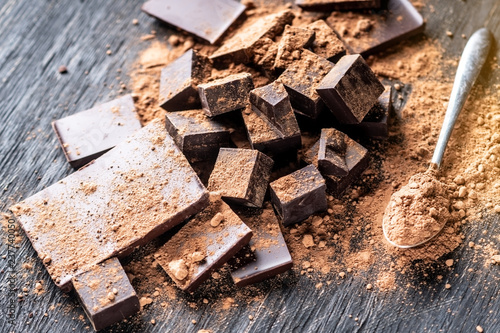 Pieces of dark bitter chocolate with cocoa powder on dark wooden background. Concept of confectionery ingredients