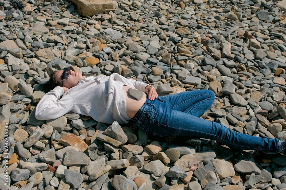 Woman in sunglasses lying on a stone beach in the fall. Stone on the girl's belly. Woman in sunglasses lying on a stone beach in the fall. Stone on the girl's belly.
