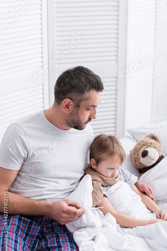 handsome father taking care of sick son in bedroom and checking his temperature with thermometer