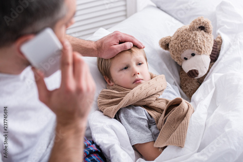 high angle view of daddy talking by smartphone and touching sick son forehead in bedroom photo