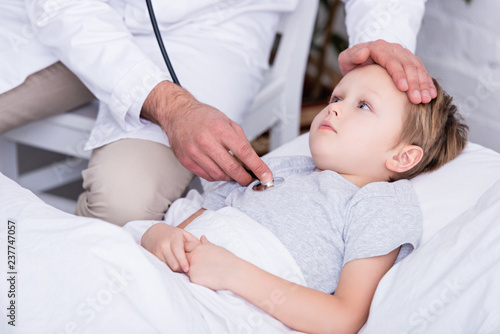 cropped image of pediatrist in white coat examining sick boy with stethoscope and touching forehead