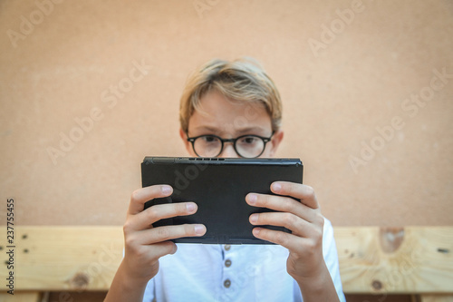 Boy on wooden bench in garden, playing absorbed with his console. Teenager engaged in favorite pastime. face with glasses behind a video game console. Employee videogame. Modern life in the open air
