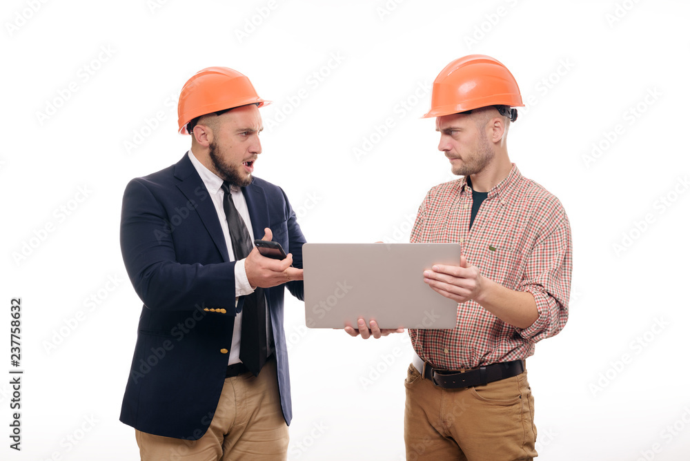 Portrait of two builders in protective orange helmets standing on white isolated background and looking at laptop display. Discuss construction project Portrait of two builders in protective orange helmets standing on white isolated background and looking at laptop display. Discuss construction project