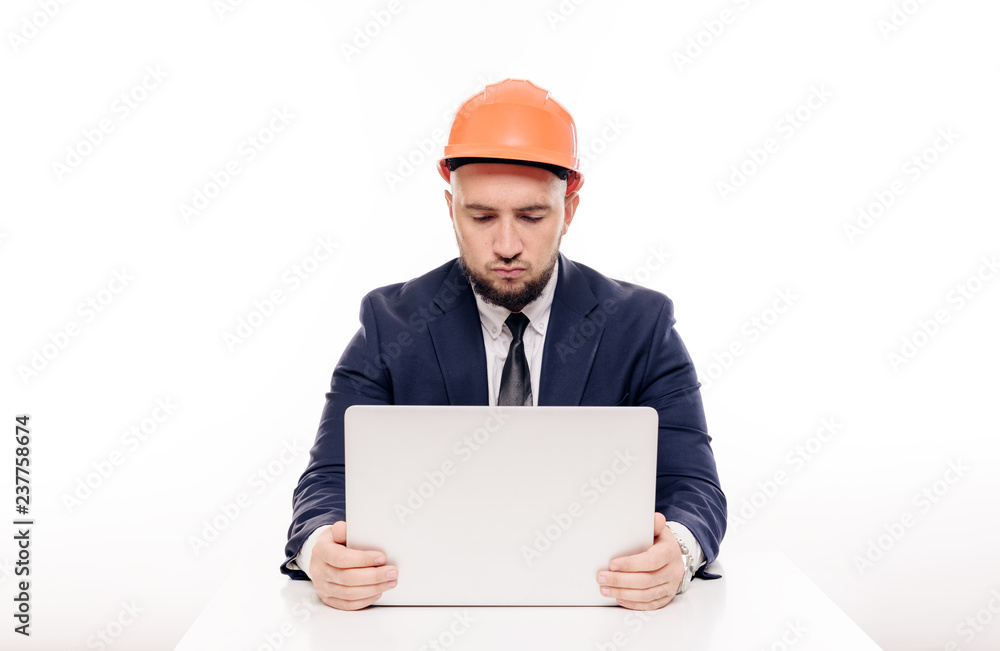 A tired constructor businessman in an orange helmet looks at the laptop screen and studies the construction project. Sits at the table and drinks coffee, nervous
