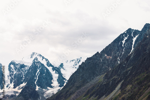 Snowy mountain top behind rocky mountain with trees under overcast sky. Rocky ridge in mist. Atmospheric minimalistic landscape of majestic nature.