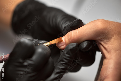 Close-up details shot of beautician hands in black rubber gloves applying with brush transparent gel polish on woman fingernail at beauty salon. Healthcare, beauty cosmetics, spa procedure concept.