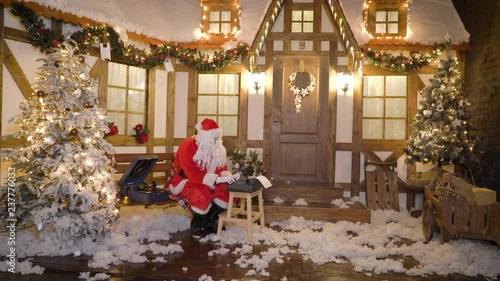 Santa Claus is printing the letter by typewriter, sits outside the house between christmas trees, drinks milk, listens music on vinyl by gramophone, preparing for Christmas