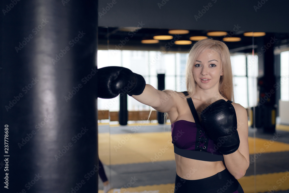 beautiful blonde girl in boxing gloves pushes the bag on a black background.Young woman training punch boxing gloves for punching bag. Girl making Strong kick.