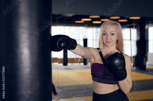 beautiful blonde girl in boxing gloves pushes the bag on a black background.Young woman training punch boxing gloves for punching bag. Girl making Strong kick. © Delete