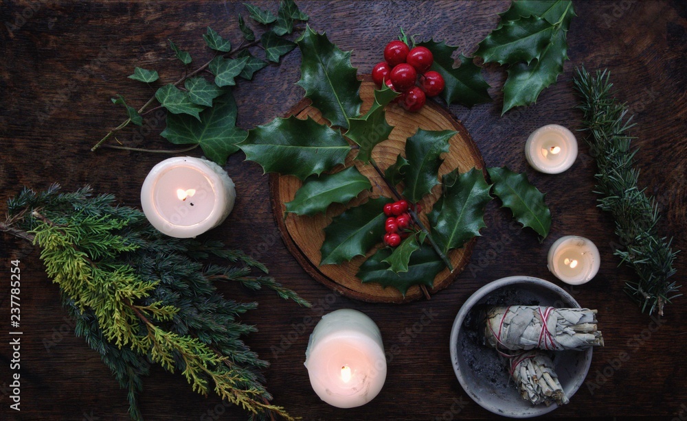 Yule winter solstice (Christmas) themed flat lay with branch of holly plant  on a dark wooden table. Sage smudge sticks, white lit burning candles,  dried evergreens, ivy branches, berries in background Photos |