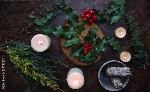 Yule winter solstice (Christmas) themed flat lay with branch of holly plant on a dark wooden table. Sage smudge sticks, white lit burning candles, dried evergreens, ivy branches, berries in background photo