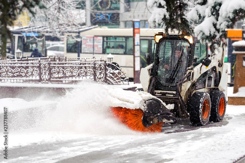 City service cleaning snow , a small tractor with a rotating brush clears a  road in the city park from the fresh fallen snow on  winter day, in the background public transport