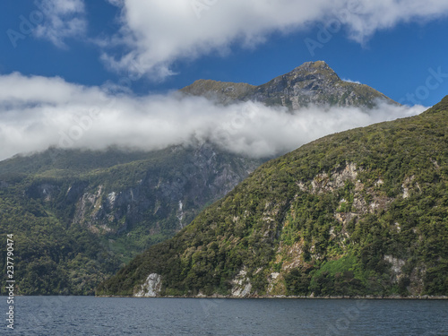 A break in the clouds reveals the rugged peaks of rocky mountainsides rising right from the water on a cruise through New Zealand’s Doubtful Sound