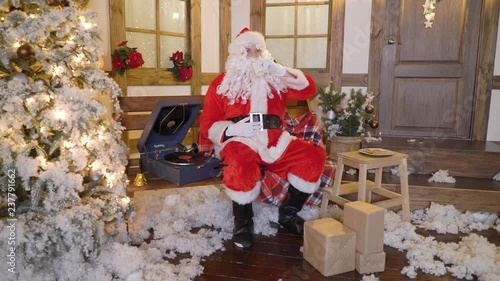 Santa Claus is packing gifts or presents in bag, sits outside the house between christmas trees, drinks milk, listens christmas songs on vinyl by gramophone, preparing for Christmas