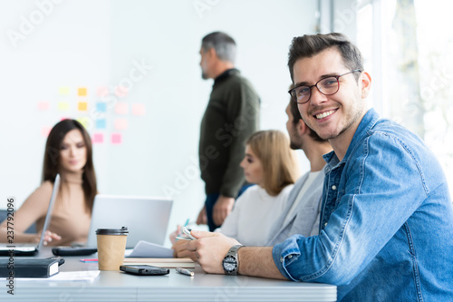 Enjoying his work. Handsome young man looking at camera and smiling while his colleagues working in the background.