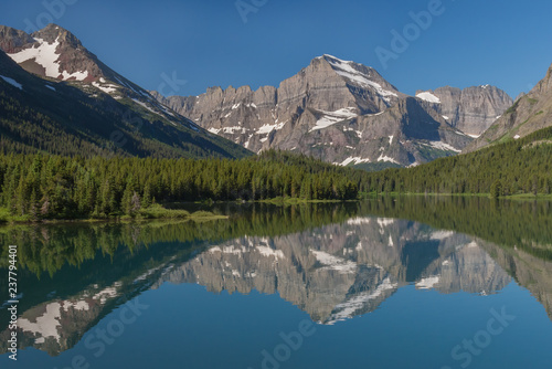 Reflections of the rugged mountains towering above the forested shores can be seen in Swiftcurrent Lake on a beautiful summer day in Glacier National Park