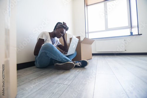 Sad young african woman sitting on floor near moving boxes and looking on screen of laptop. Young woman moving to new home. © F8  \ Suport Ukraine