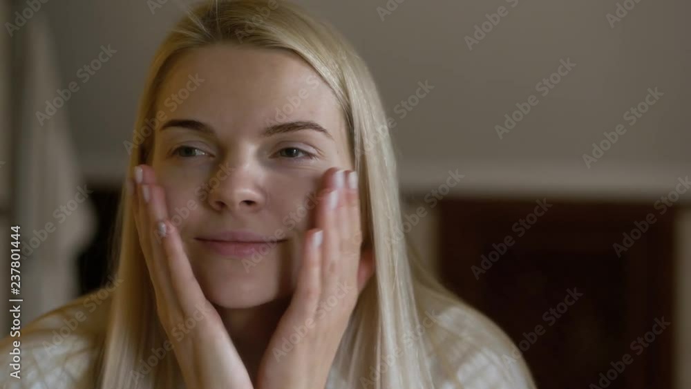 Young adorable woman in white towel turban doing her daily beauty care cosmetic routine, examines her face at mirror in the bathroom and smiling. Young adorable woman in white towel turban doing her daily beauty care cosmetic routine, examines her face at mirror in the bathroom and smiling.
