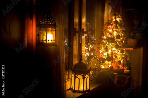 desk of free space and lamp with xmas tree in home . Christmas lantern in selective focus near window with holiday tree full of colored toys and lights.