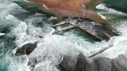 Wide aerial shot of waves and powerful ocean swell crashing over Monavale Rock Pool on a turbulent spring day. 50FPS SLOW MOTION photo