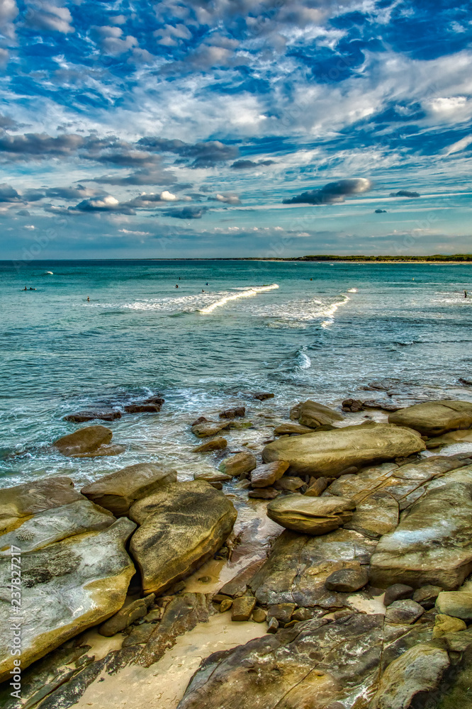 Beach seascape with aged old rocky surface, soft waves and clouds surfers in the distance