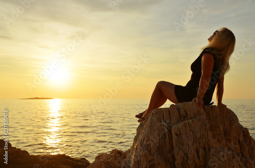 A woman sitting on a cliff is watching the sunset over the sea.