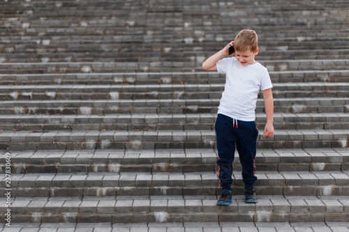 A boy in sports clothes down the stairs and talking on the phone outdoors