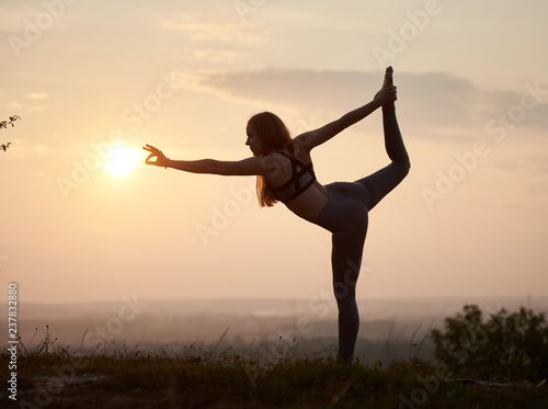 Silhouette of slim strong muscular girl standing on one leg doing complicated yoga exercises outdoors at dawn on copy space background of light pink summer sky and blurred misty horizon. Narajasana