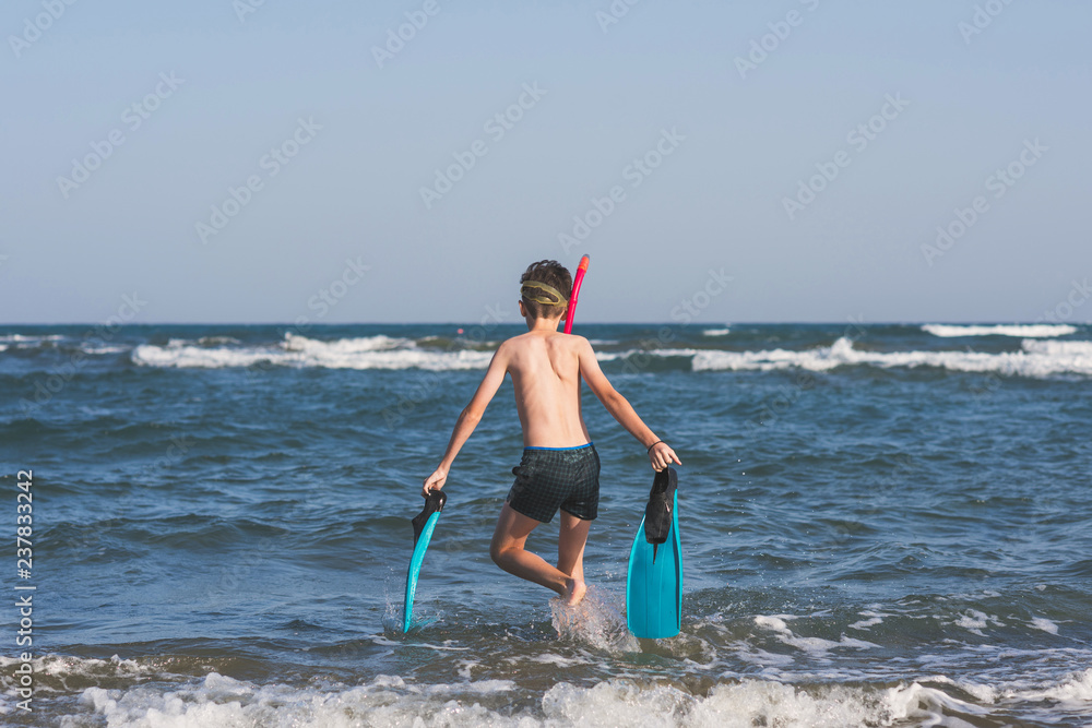 Fototapeta premium Happy teen boy in the swim flippers and snorkeling mask running оn the wave of the sea during summer vacation in the tropical resort town
