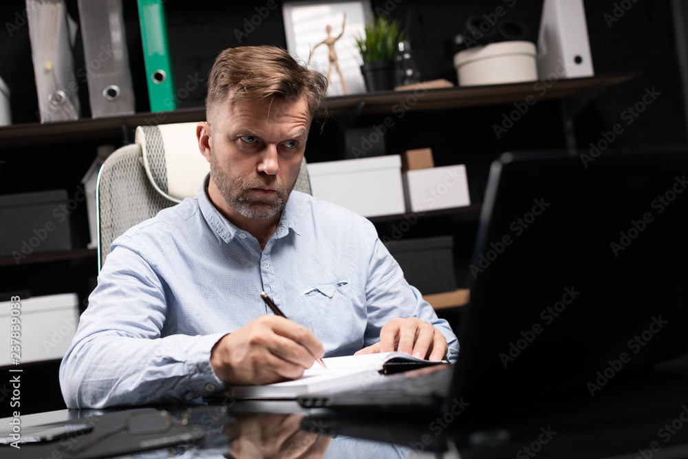 young man sitting at computer table and makes notes in diary