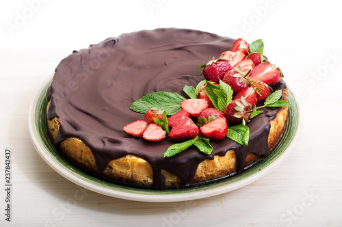 Cheesecake (homemade cake), poured with chocolate icing, with strawberry berries in a plate (side view). Sweets on a white wooden board. Organic product.