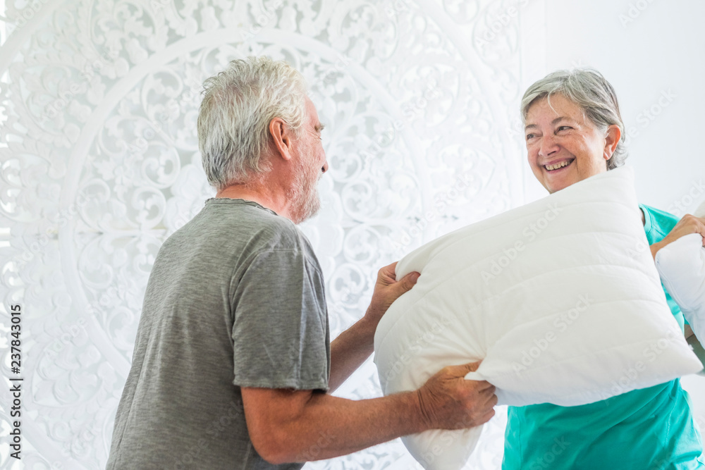 Aged senior adult caucasian couple have fun making war with pillows at home in the bedroom at morning - white bright room and house activity for cheerful old people Aged senior adult caucasian couple have fun making war with pillows at home in the bedroom at morning - white bright room and house activity for cheerful old people