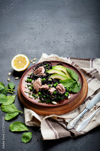 Pasta with cuttlefish ink (black noodles) with tuna pieces, spinach leaves, avocado slices, green peas, onions and sunflower seeds on a clay plate. An original pasta recipe. Useful and tasty dish